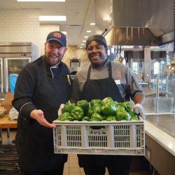 Two Tiger Dining chefs smiling and holding a crate of green bell peppers in Wellness Kitchen..